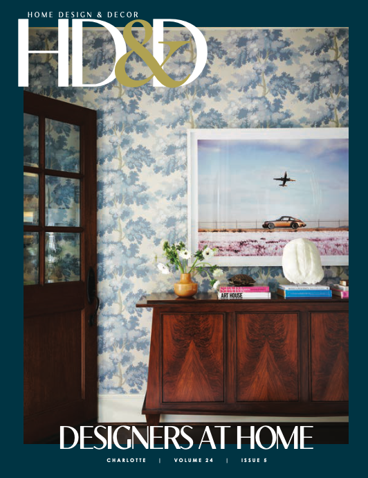 Magazine cover featuring an interior scene with a wooden sideboard, a framed airplane and car photograph, a vase with flowers, and decorative objects against blue floral wallpaper.
