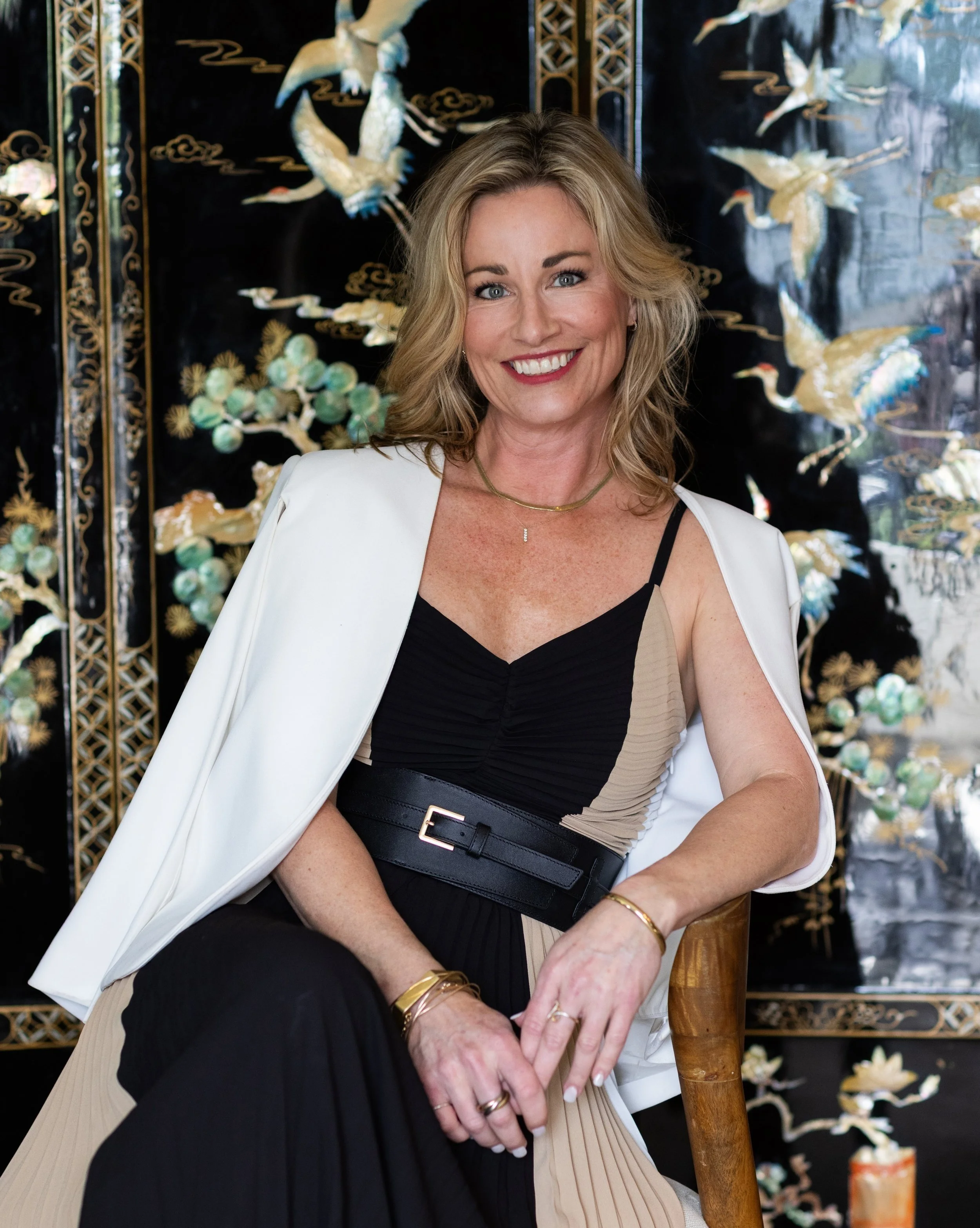 A smiling woman with blonde hair sitting in front of an Asian-inspired folding screen with cranes and floral designs, wearing a black and beige pleated dress with a wide black belt and a white blazer draped over her shoulders.