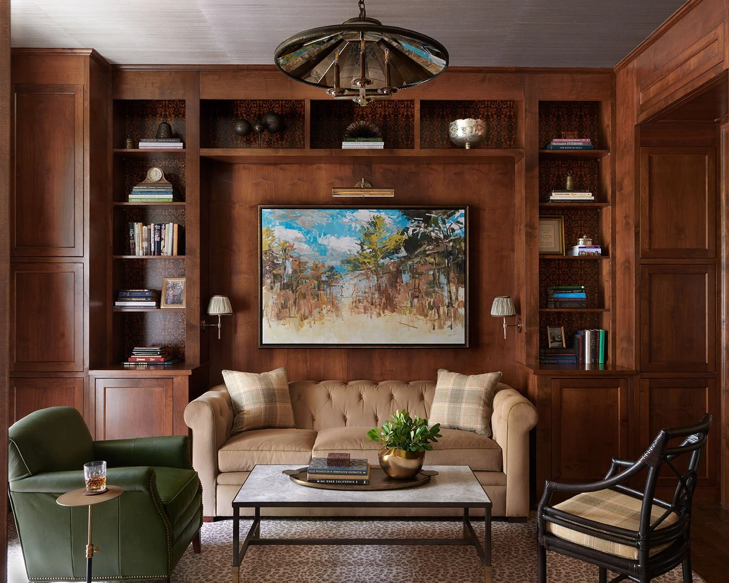 Living room with wood-paneled walls, a beige tufted sofa, a green armchair with a drink, and a black chair. A coffee table with books and a plant is in front. A large framed painting hangs on the wall, flanked by two small wall sconces. Built-in bookshelves with books and decorative items frame the seating area. Overhead, a unique chandelier hangs.